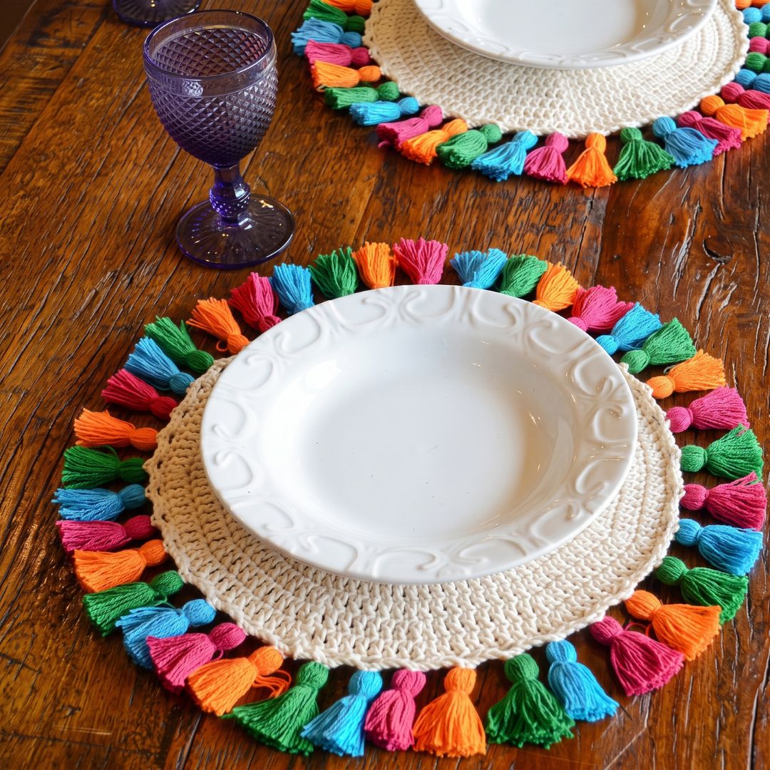 A close-up of a place setting on a rustic wooden table, featuring a round cream-colored crocheted placemat with vibrant multicolored tassels, a white ceramic plate, and a purple glass goblet.