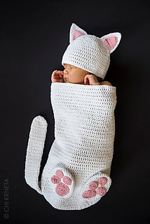 Newborn baby in a handmade crochet white cat cocoon with cat ear hat, tail and pink paw details