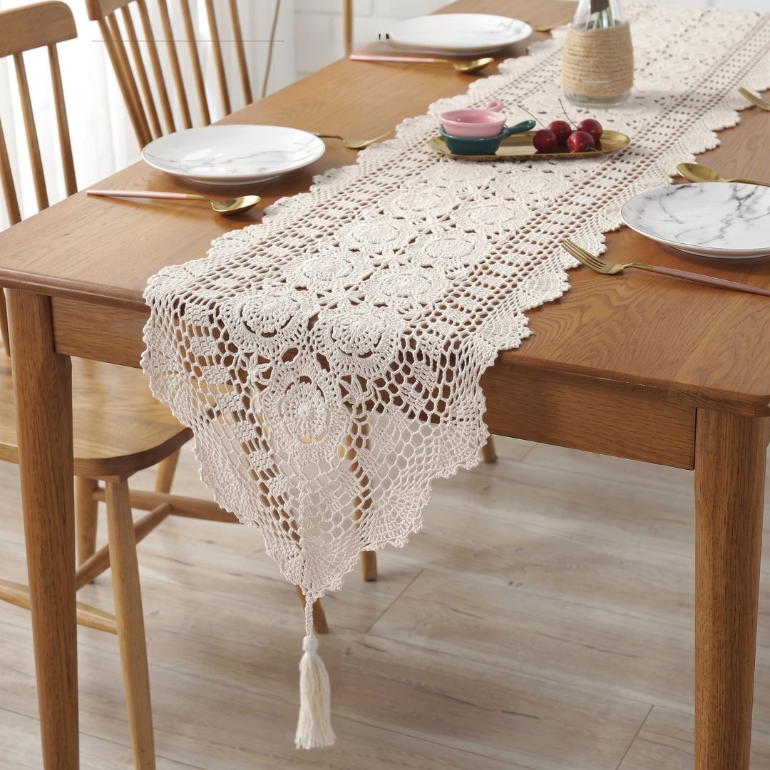A cream-colored crocheted table runner with an intricate lace pattern and a tassel at the end, draped over a wooden dining table set with plates and cutlery.