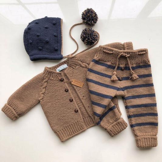 A baby knit outfit set laid flat on a light surface, including a tan cardigan, matching striped pants, a navy bonnet, and a pom-pom headband.
