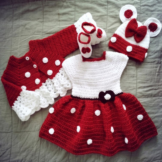 A handmade crocheted baby outfit set laid flat on a gray quilted blanket. The set includes a red and white dress with white polka dots, a matching red and white cardigan, a white and red bootie, and a white and red mouse-ear hat with a red bow.